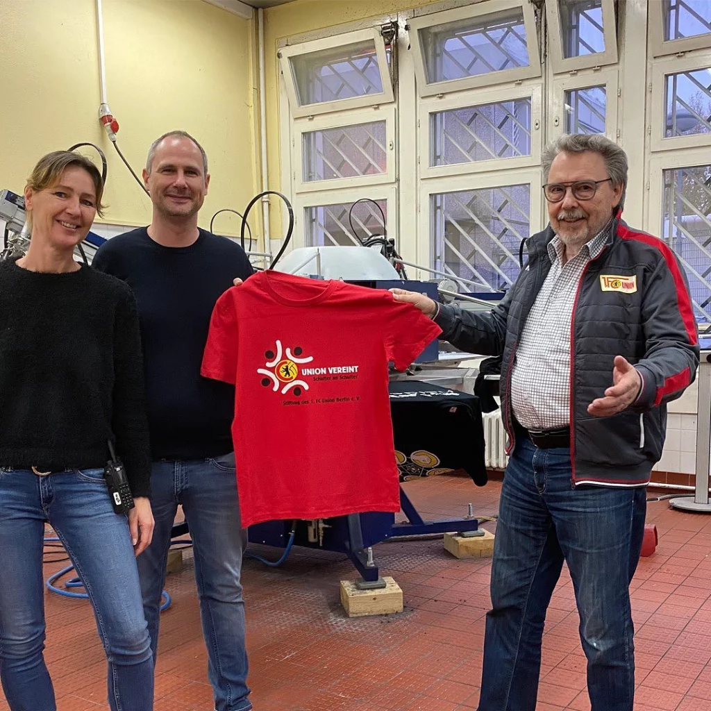 Das Team von BoOH! mit einem bärtigen, älterem Mann von 1. FC Union. Gemeinsam halten sie ein rotes T-Shirt hoch
