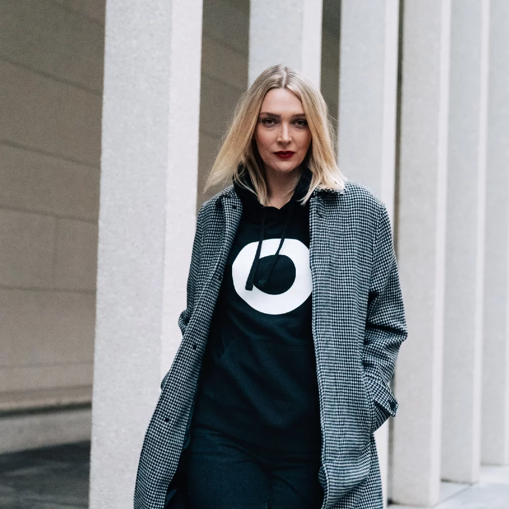 Ein blondes Model trägt einen schwarzen Hoodie mit dem Kampagnen Icon vom Weissen Ring. Darüber ein grauer Mantel.